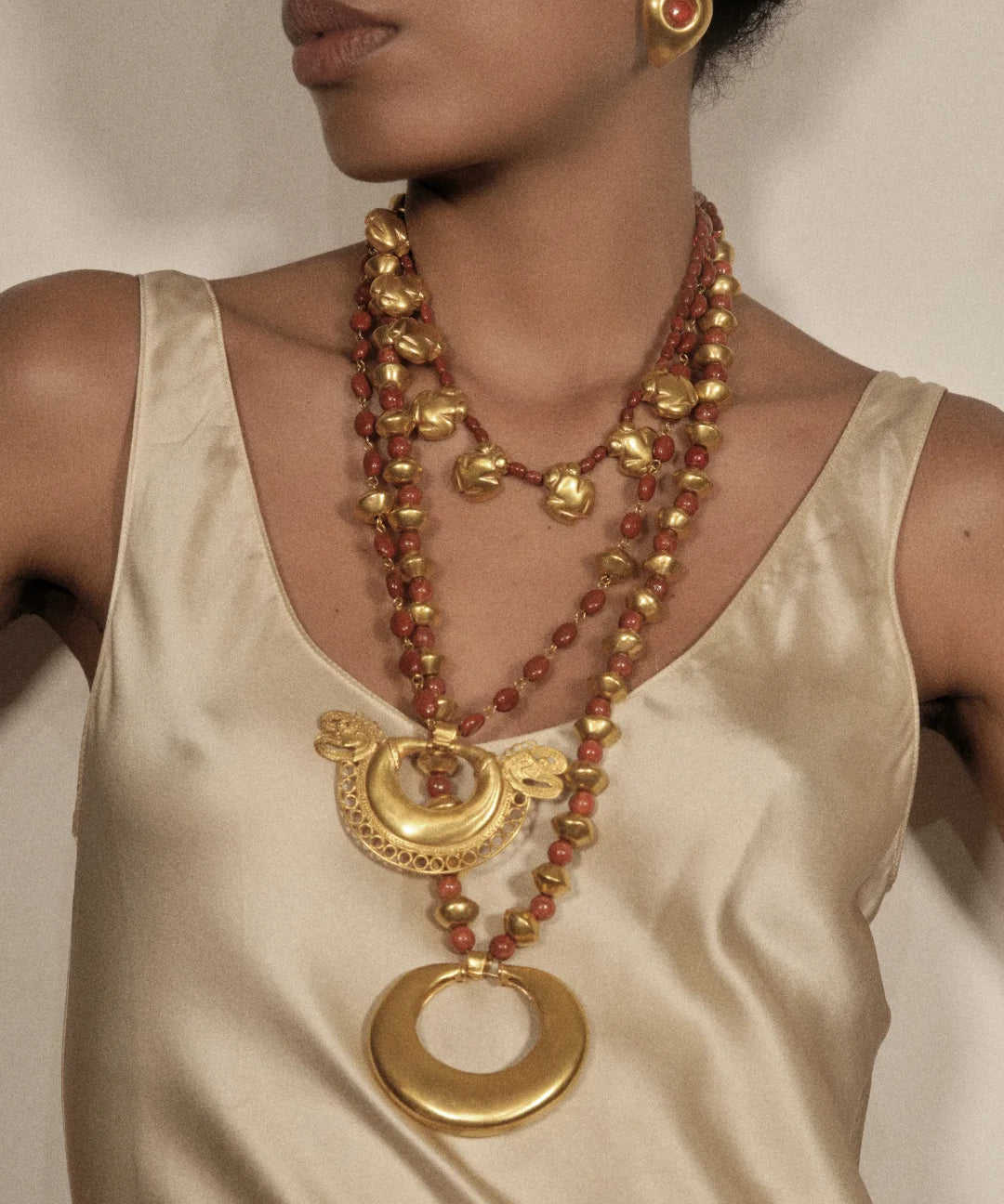 A close-up of a model wearing a multi-strand necklace featuring gold-toned metal elements and reddish-brown beads, paired with a cream satin top.
