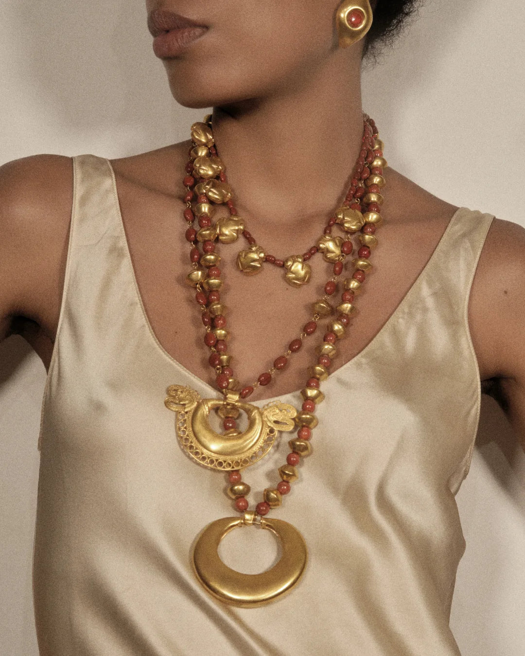 A close-up of a model wearing a multi-strand necklace featuring gold-toned metal elements and reddish-brown beads, paired with a cream satin top.
