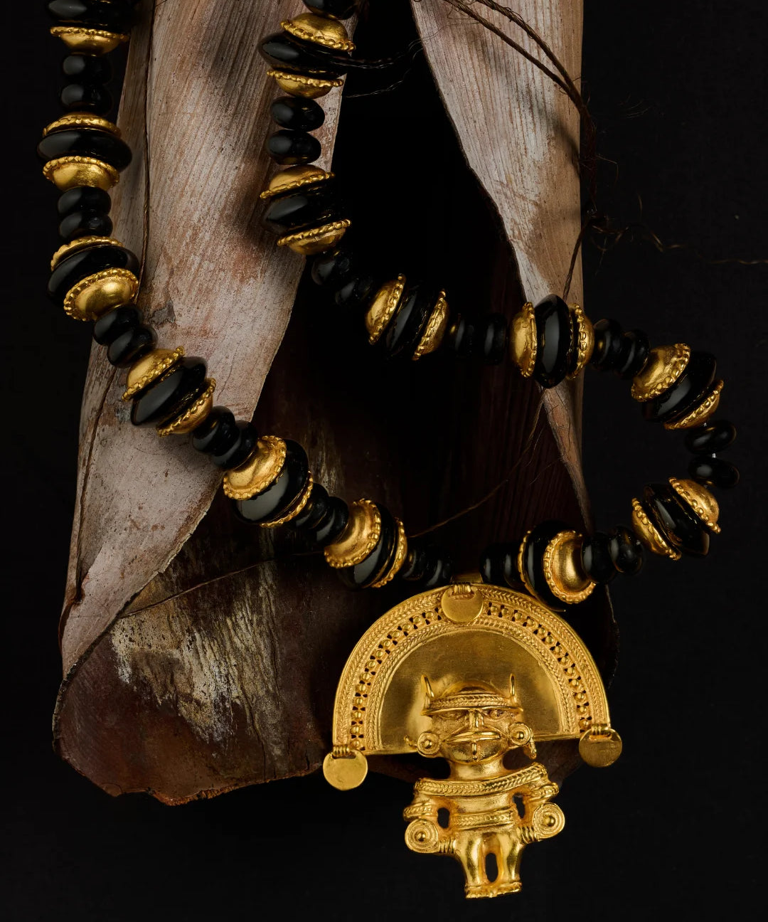 A beaded necklace with alternating black and gold beads, featuring a prominent gold pendant shaped like a stylized figure under a semi-circular arch, displayed against a dark background with dried leaves.