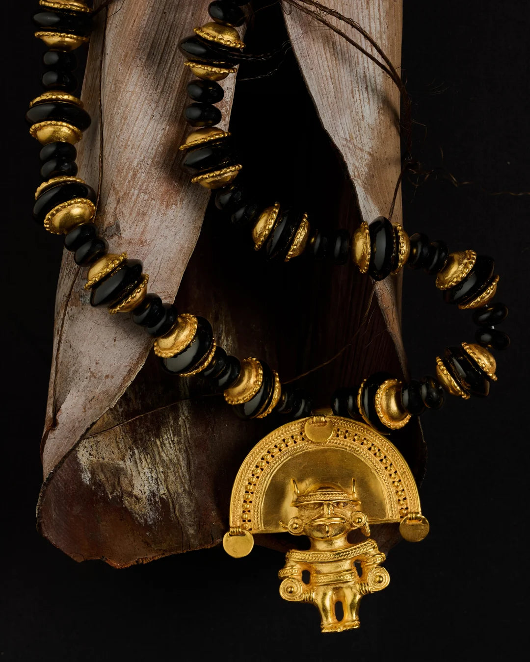 A beaded necklace with alternating black and gold beads, featuring a prominent gold pendant shaped like a stylized figure under a semi-circular arch, displayed against a dark background with dried leaves.