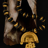 A beaded necklace with alternating black and gold beads, featuring a prominent gold pendant shaped like a stylized figure under a semi-circular arch, displayed against a dark background with dried leaves.