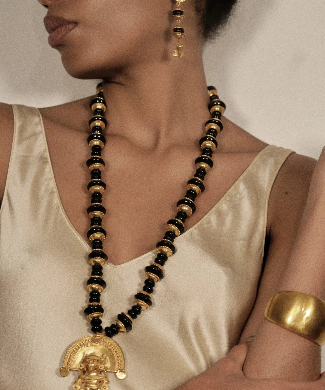 A close-up of a model wearing a long black and gold beaded necklace with a gold pendant, a matching gold earring, and a gold cuff bracelet, styled with a cream satin top.