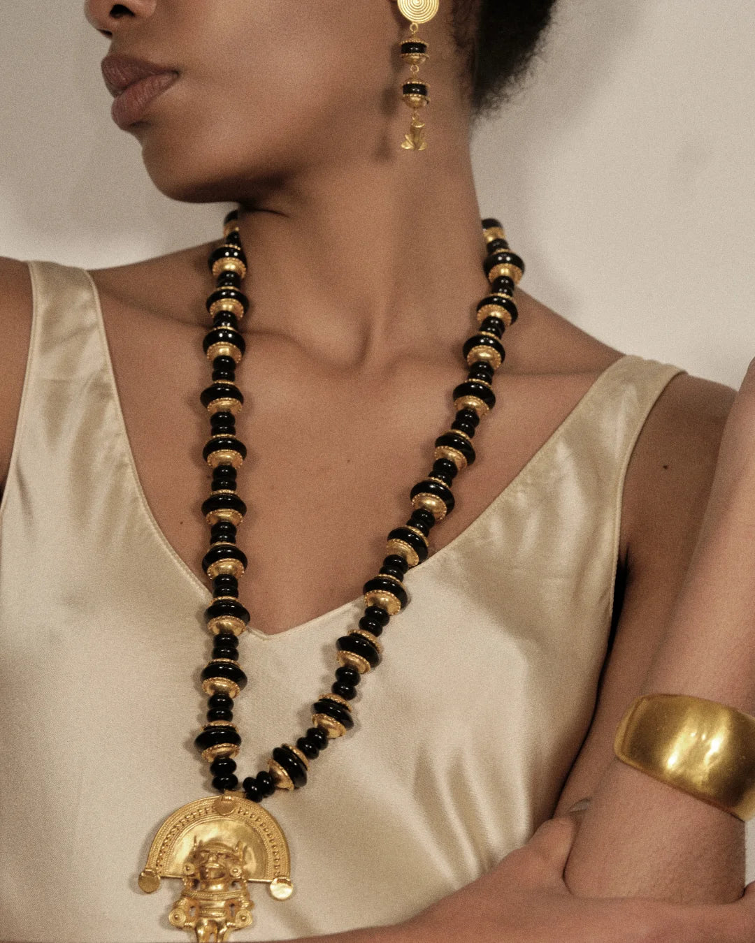 A close-up of a model wearing a long black and gold beaded necklace with a gold pendant, a matching gold earring, and a gold cuff bracelet, styled with a cream satin top.