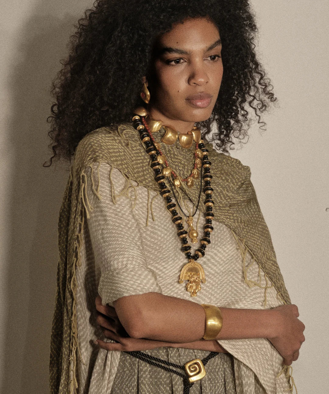 A model with voluminous dark curly hair is shown from the chest up, wearing layered gold and black beaded necklaces, a chunky gold collar, a gold cuff bracelet, and a textured beige and olive green fringed shawl over a patterned top, set against a plain beige wall.