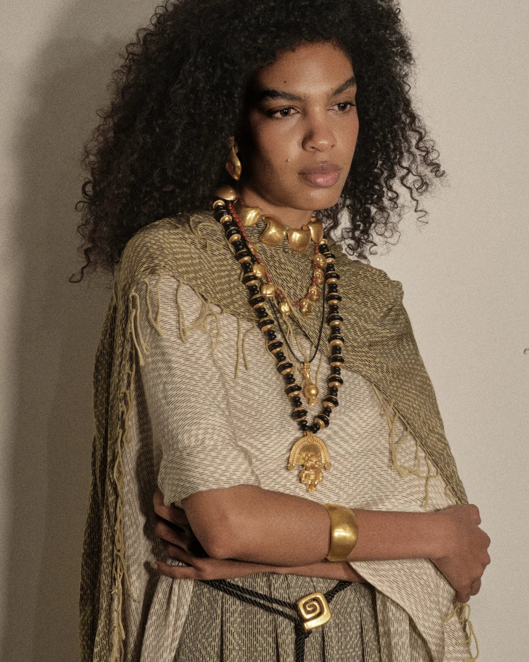 A model with voluminous dark curly hair is shown from the chest up, wearing layered gold and black beaded necklaces, a chunky gold collar, a gold cuff bracelet, and a textured beige and olive green fringed shawl over a patterned top, set against a plain beige wall.