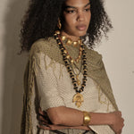 A model with voluminous dark curly hair is shown from the chest up, wearing layered gold and black beaded necklaces, a chunky gold collar, a gold cuff bracelet, and a textured beige and olive green fringed shawl over a patterned top, set against a plain beige wall.