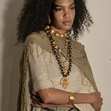 A model with voluminous dark curly hair is shown from the chest up, wearing layered gold and black beaded necklaces, a chunky gold collar, a gold cuff bracelet, and a textured beige and olive green fringed shawl over a patterned top, set against a plain beige wall.
