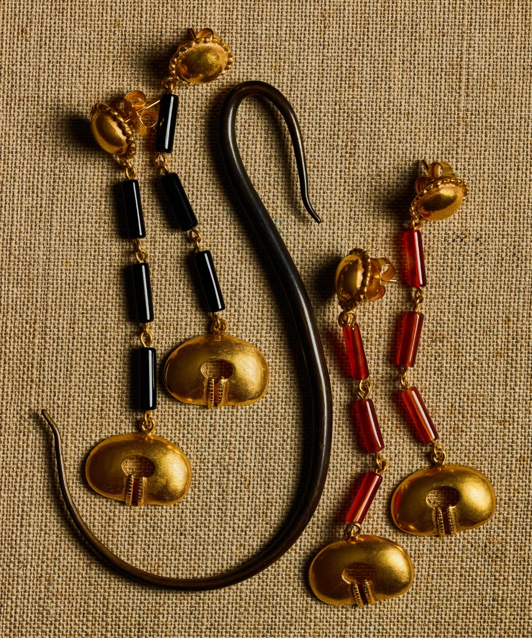 A pair of ornate gold-toned earrings with black cylindrical beads and red cylindrical beads, displayed on a textured beige fabric background next to a curved black object.