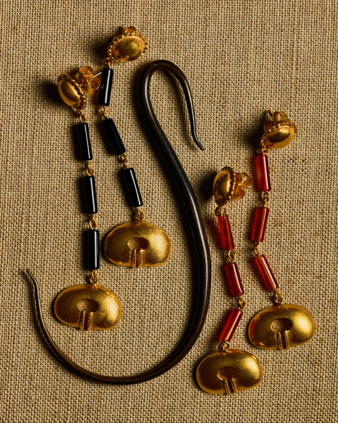 A pair of ornate gold-toned earrings with black cylindrical beads and red cylindrical beads, displayed on a textured beige fabric background next to a curved black object.