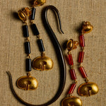 A pair of ornate gold-toned earrings with black cylindrical beads and red cylindrical beads, displayed on a textured beige fabric background next to a curved black object.