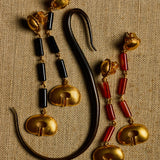 A pair of ornate gold-toned earrings with black cylindrical beads and red cylindrical beads, displayed on a textured beige fabric background next to a curved black object.