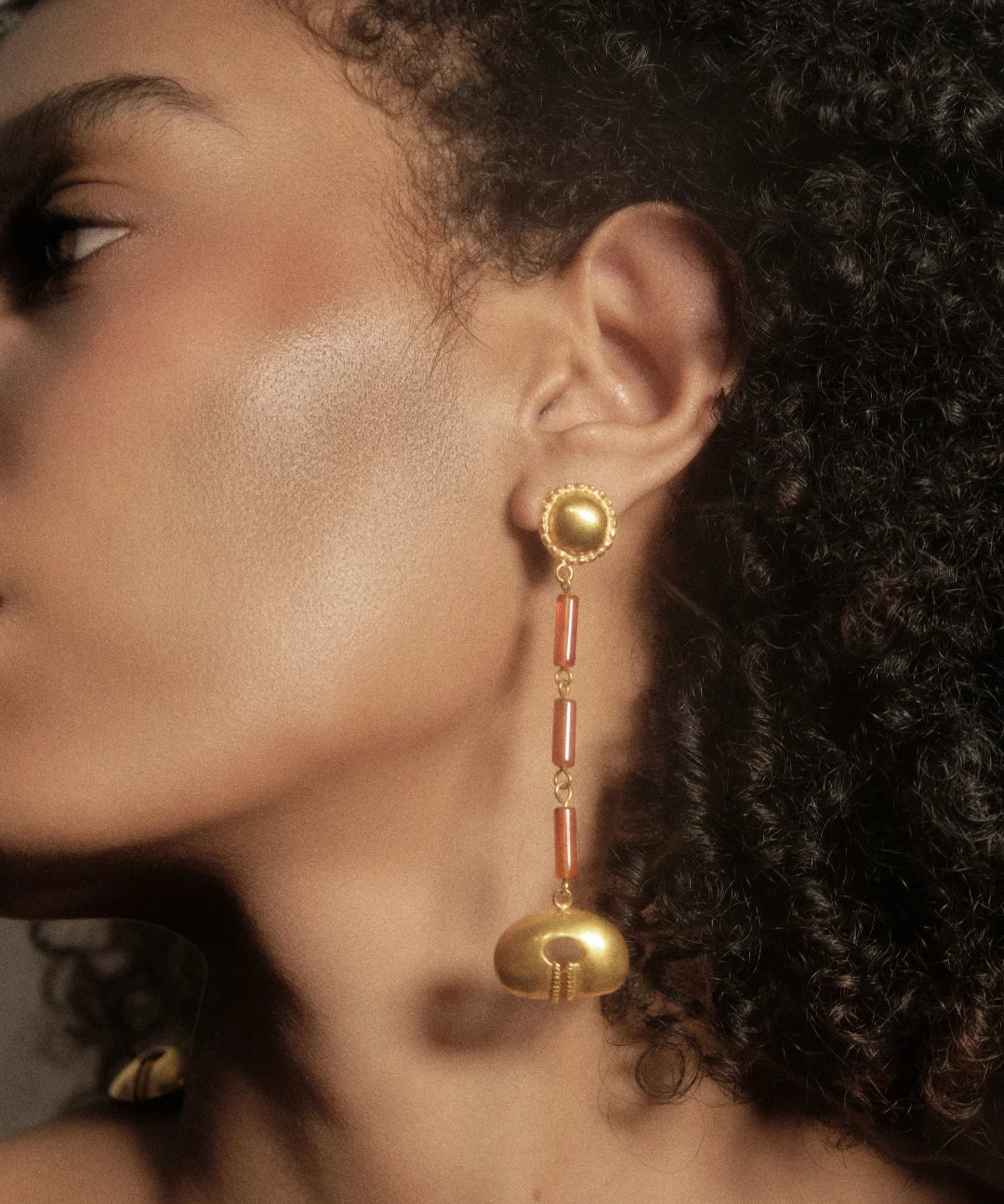 A close-up side profile of a woman wearing a long, ornate gold dangle earring with red cylindrical accents and a gold bell-shaped drop, set against dark curly hair.