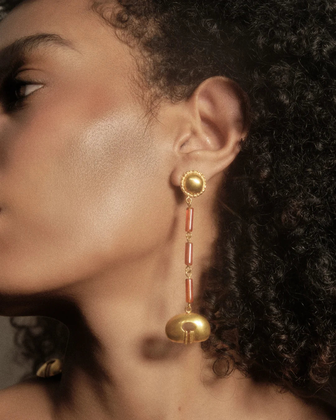 A close-up side profile of a woman wearing a long, ornate gold dangle earring with red cylindrical accents and a gold bell-shaped drop, set against dark curly hair.