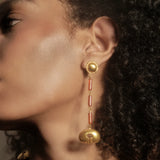 A close-up side profile of a woman wearing a long, ornate gold dangle earring with red cylindrical accents and a gold bell-shaped drop, set against dark curly hair.