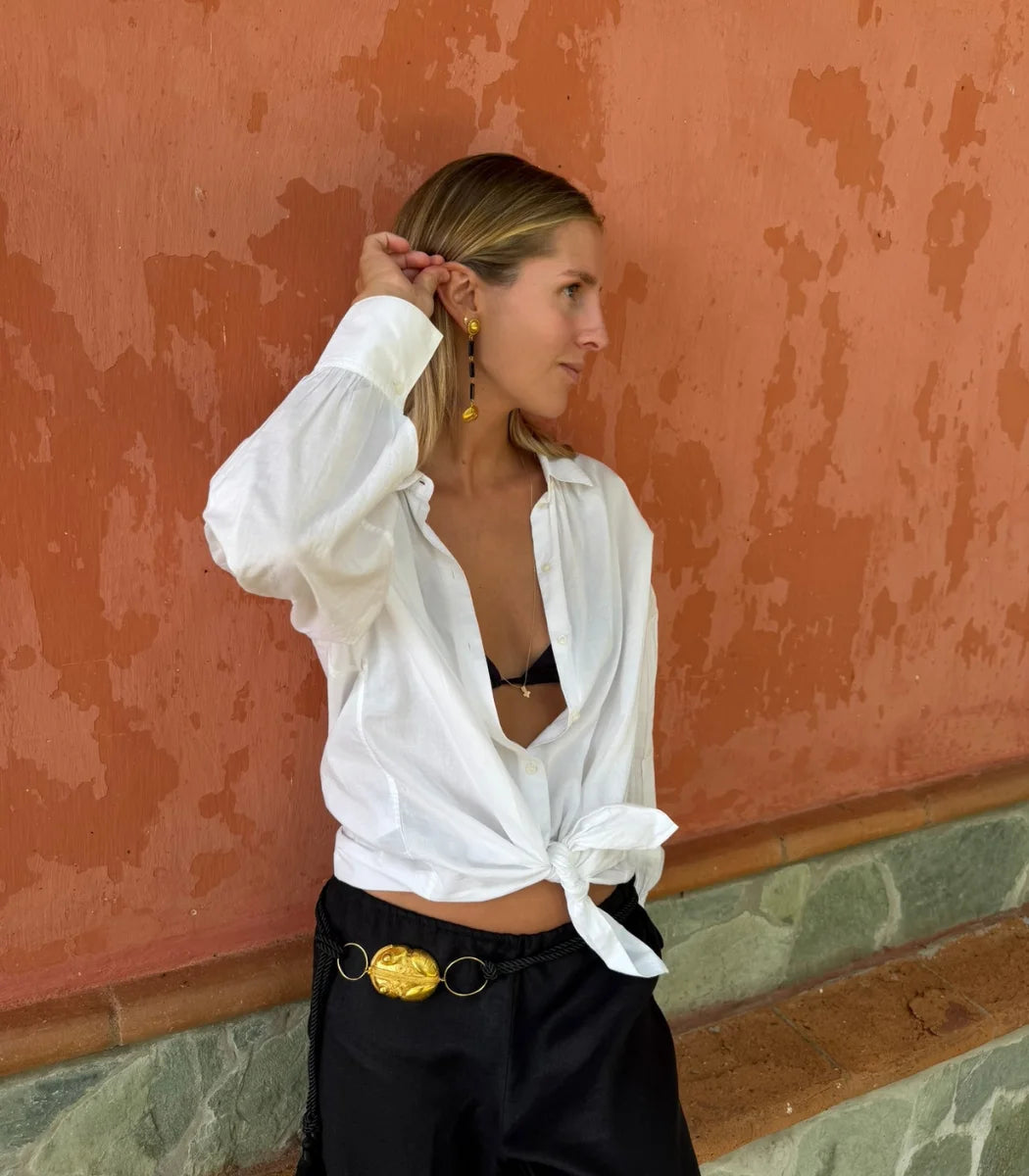 A woman wearing a white long-sleeve button-up shirt tied at the waist and black pants with a gold belt buckle, posing against a textured terracotta wall.