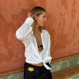 A woman wearing a white long-sleeve button-up shirt tied at the waist and black pants with a gold belt buckle, posing against a textured terracotta wall.