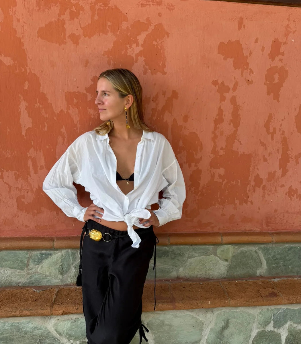 A woman posing in front of a textured terracotta wall, wearing a white long-sleeve button-down shirt tied at the waist over a black bralette, paired with black pants and a gold belt.