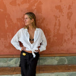 A woman posing in front of a textured terracotta wall, wearing a white long-sleeve button-down shirt tied at the waist over a black bralette, paired with black pants and a gold belt.