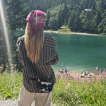 A person standing outdoors by a green lake, seen from behind, wearing a dark striped shirt, beige pants, a pink bandana, and a small crossbody bag with a colorful face design. Trees and wildflowers are visible in the background.