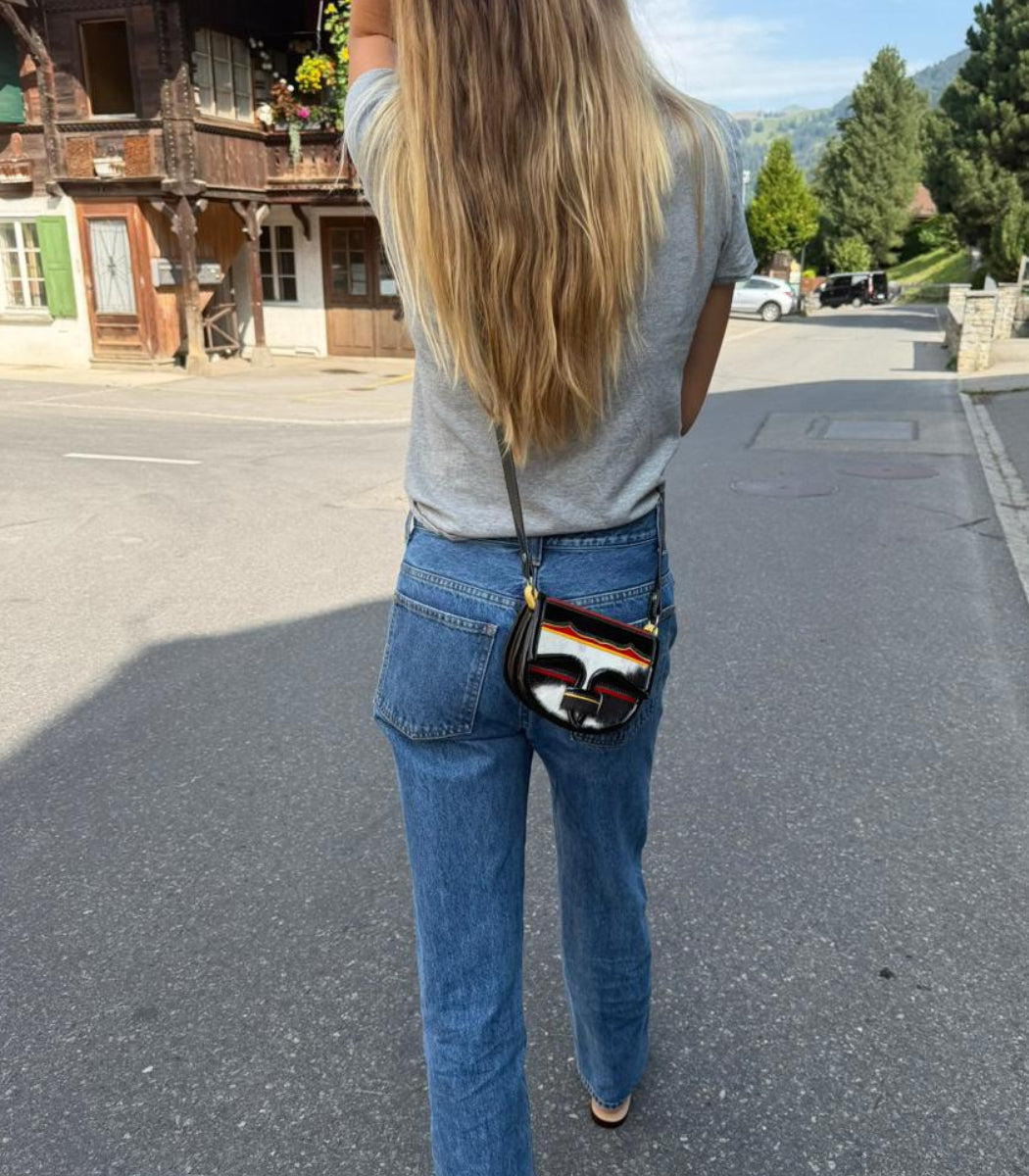 A woman viewed from behind wearing a gray t-shirt, blue jeans, and carrying a small black crossbody bag with a colorful striped pattern and a gold-tone Fendi logo on the back of the pocket.
