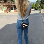 A woman viewed from behind wearing a gray t-shirt, blue jeans, and carrying a small black crossbody bag with a colorful striped pattern and a gold-tone Fendi logo on the back of the pocket.