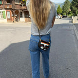 A woman viewed from behind wearing a gray t-shirt, blue jeans, and carrying a small black crossbody bag with a colorful striped pattern and a gold-tone Fendi logo on the back of the pocket.