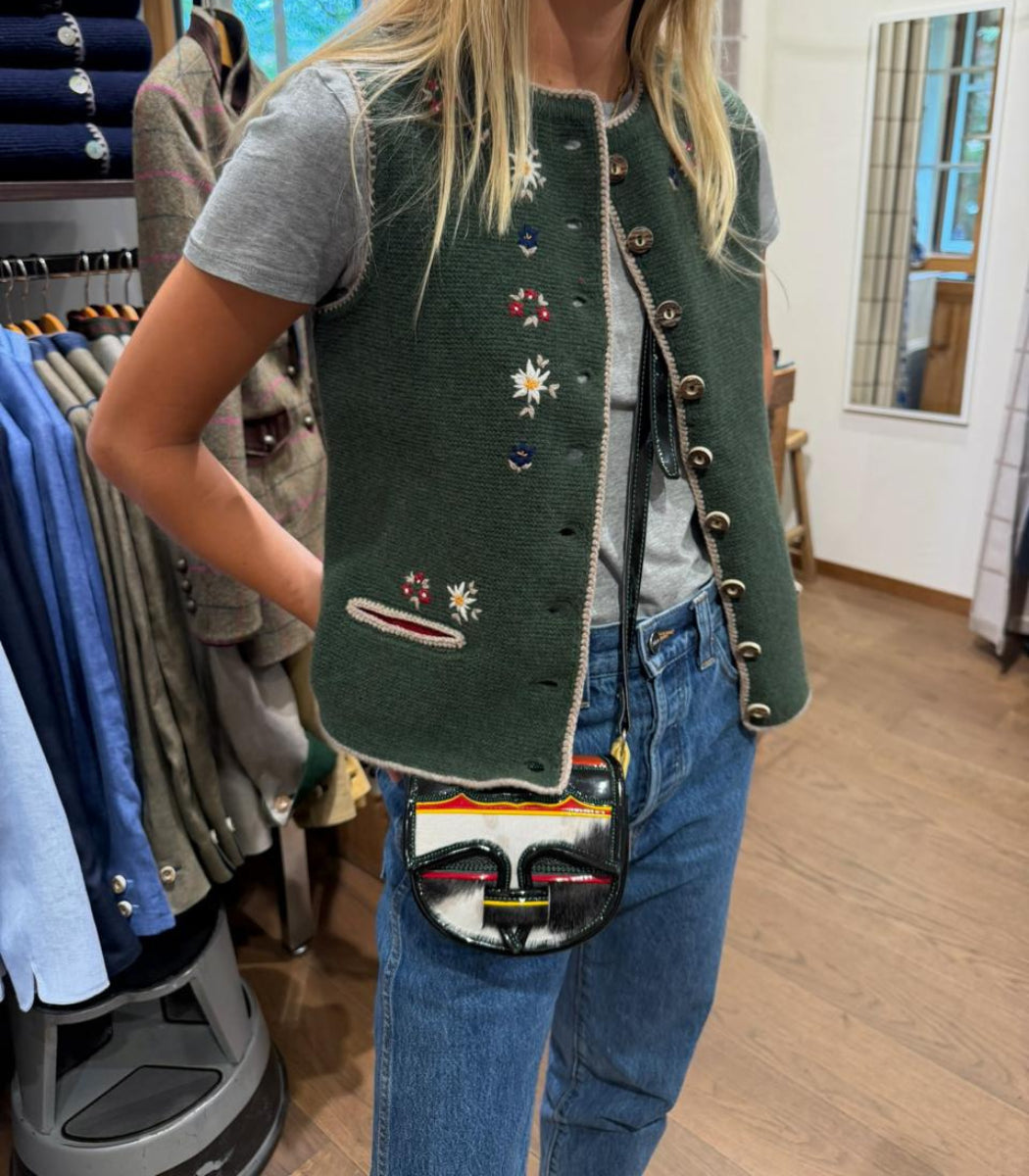 A woman wearing a dark green sleeveless vest with colorful embroidered flowers and decorative buttons, styled over a gray t-shirt and blue jeans, carrying a small black and white handbag with red, yellow, and green stripes.