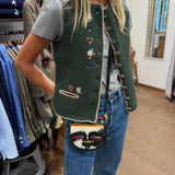 A woman wearing a dark green sleeveless vest with colorful embroidered flowers and decorative buttons, styled over a gray t-shirt and blue jeans, carrying a small black and white handbag with red, yellow, and green stripes.