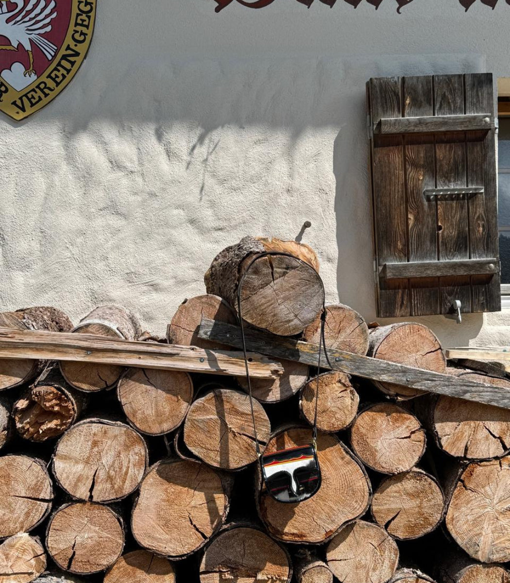 A pair of black sunglasses with orange and white stripes on the lens frame hangs from a wooden beam resting on a stacked pile of cut logs in front of a white stucco wall with a dark wooden shutter.