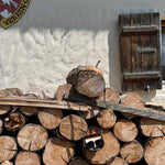 A pair of black sunglasses with orange and white stripes on the lens frame hangs from a wooden beam resting on a stacked pile of cut logs in front of a white stucco wall with a dark wooden shutter.