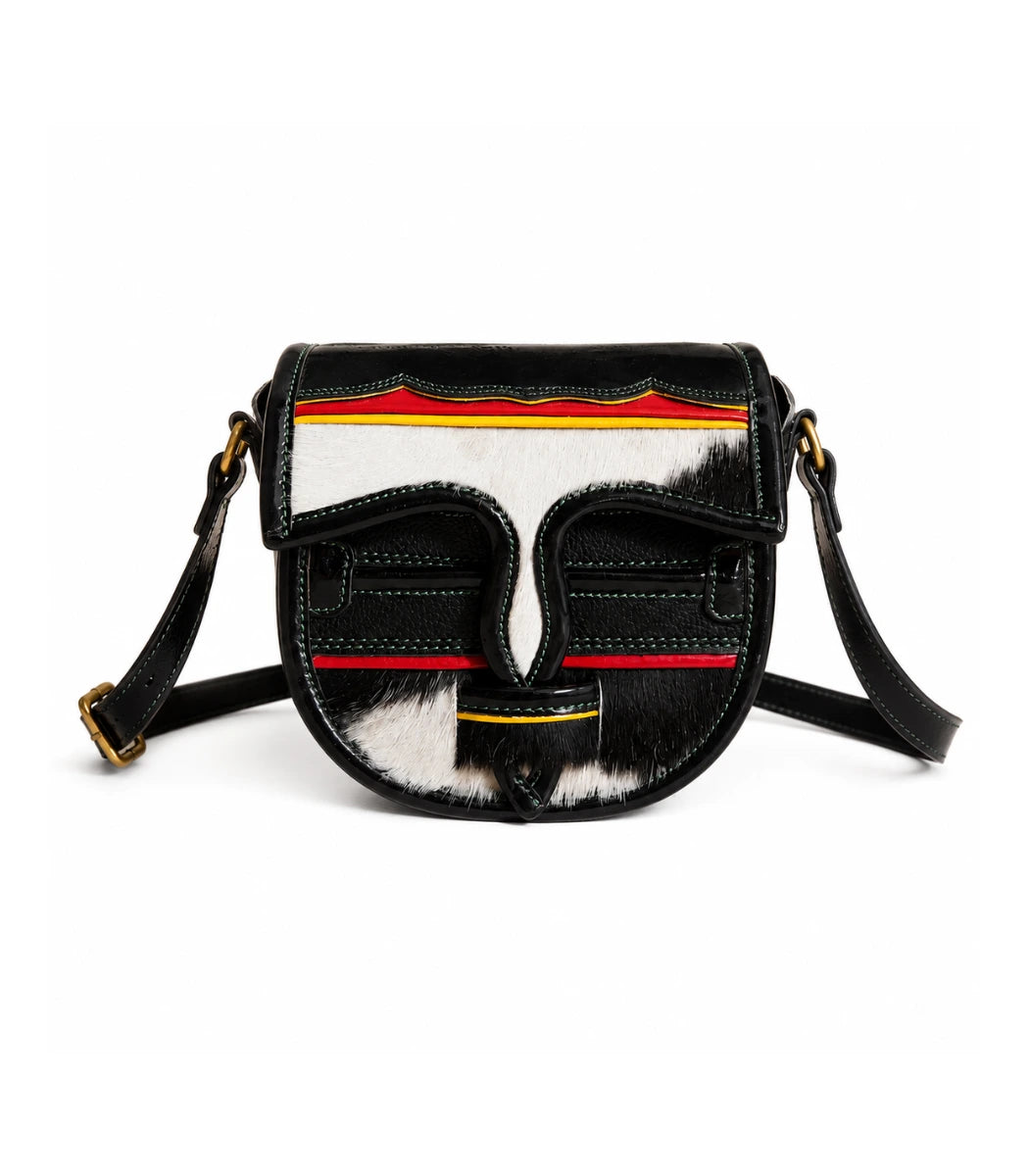 A black leather shoulder bag with a curved front featuring a stylized face design made from black, white, and brown hair-on-hide, accented with red and yellow stripes, on a white background.