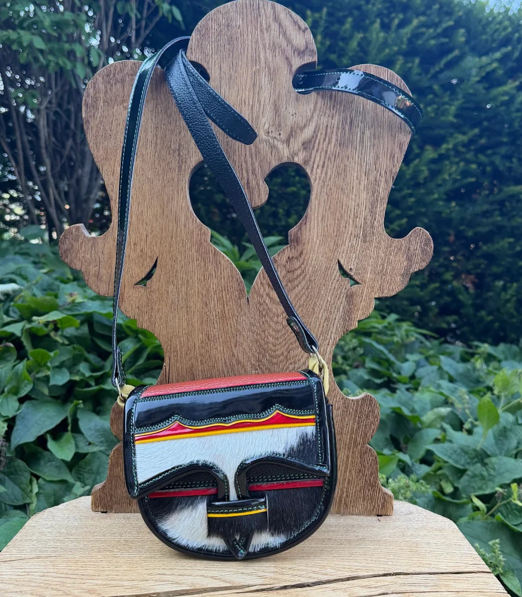 A small crossbody bag with a black patent leather body, a white, red, and yellow striped front panel, and a black-and-white cowhide flap, displayed on a wooden stand outdoors with green foliage in the background.