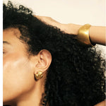 Close-up of a person with dark curly hair wearing a wide gold cuff bracelet and a matching gold stud earring against a plain light background.