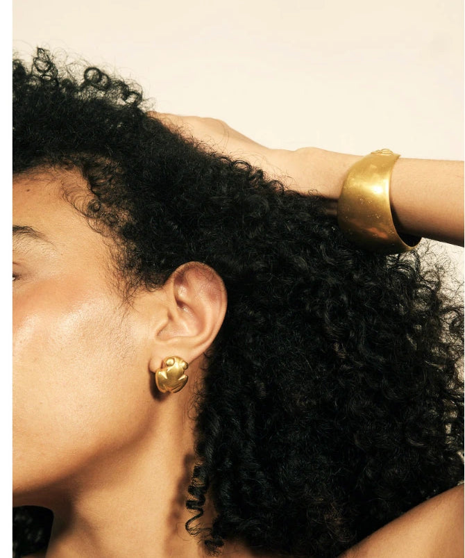 Close-up of a person with dark curly hair wearing a wide gold cuff bracelet and a matching gold stud earring against a plain light background.