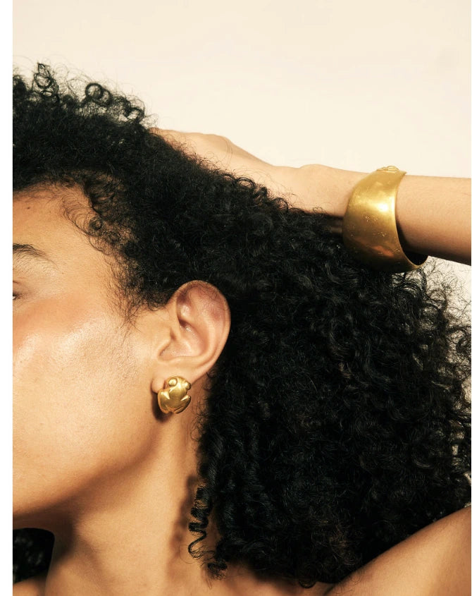 Close-up of a person with dark curly hair wearing a wide gold cuff bracelet and a matching gold stud earring against a plain light background.