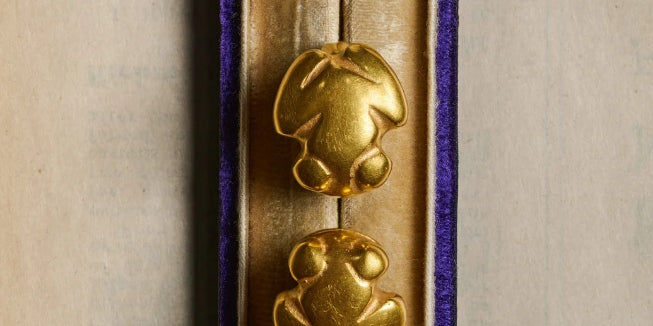 A pair of gold frog-shaped earrings displayed in an open purple velvet jewelry box on a beige background.