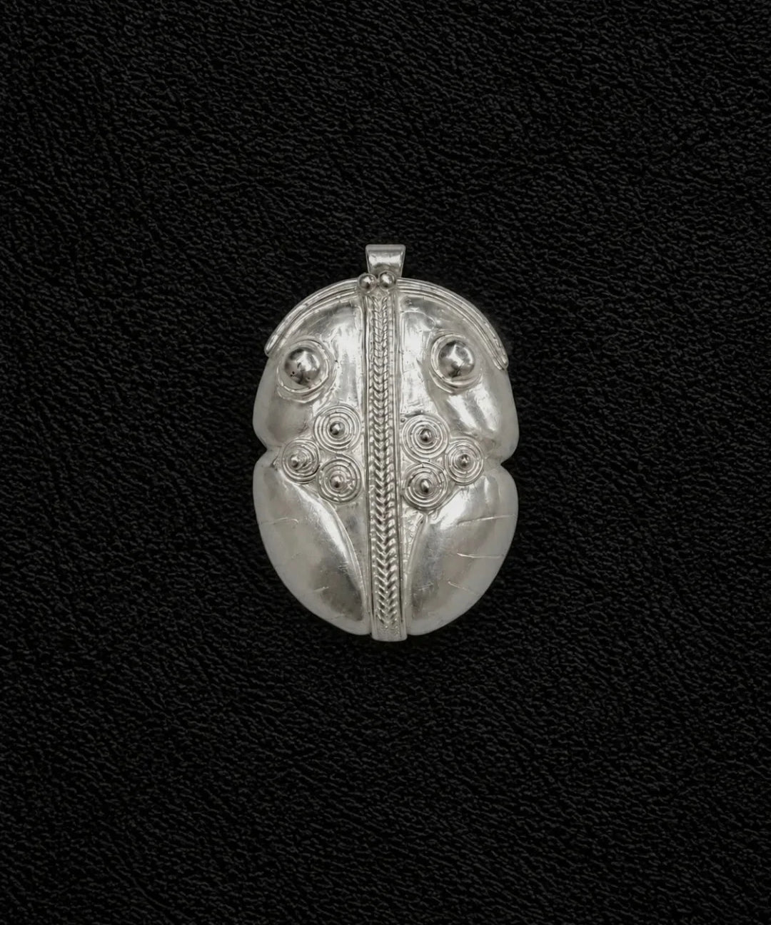 A silver pendant with intricate tribal-style engraved patterns, including a central vertical line, circular details, and leaf-like sections, displayed on a textured black background.