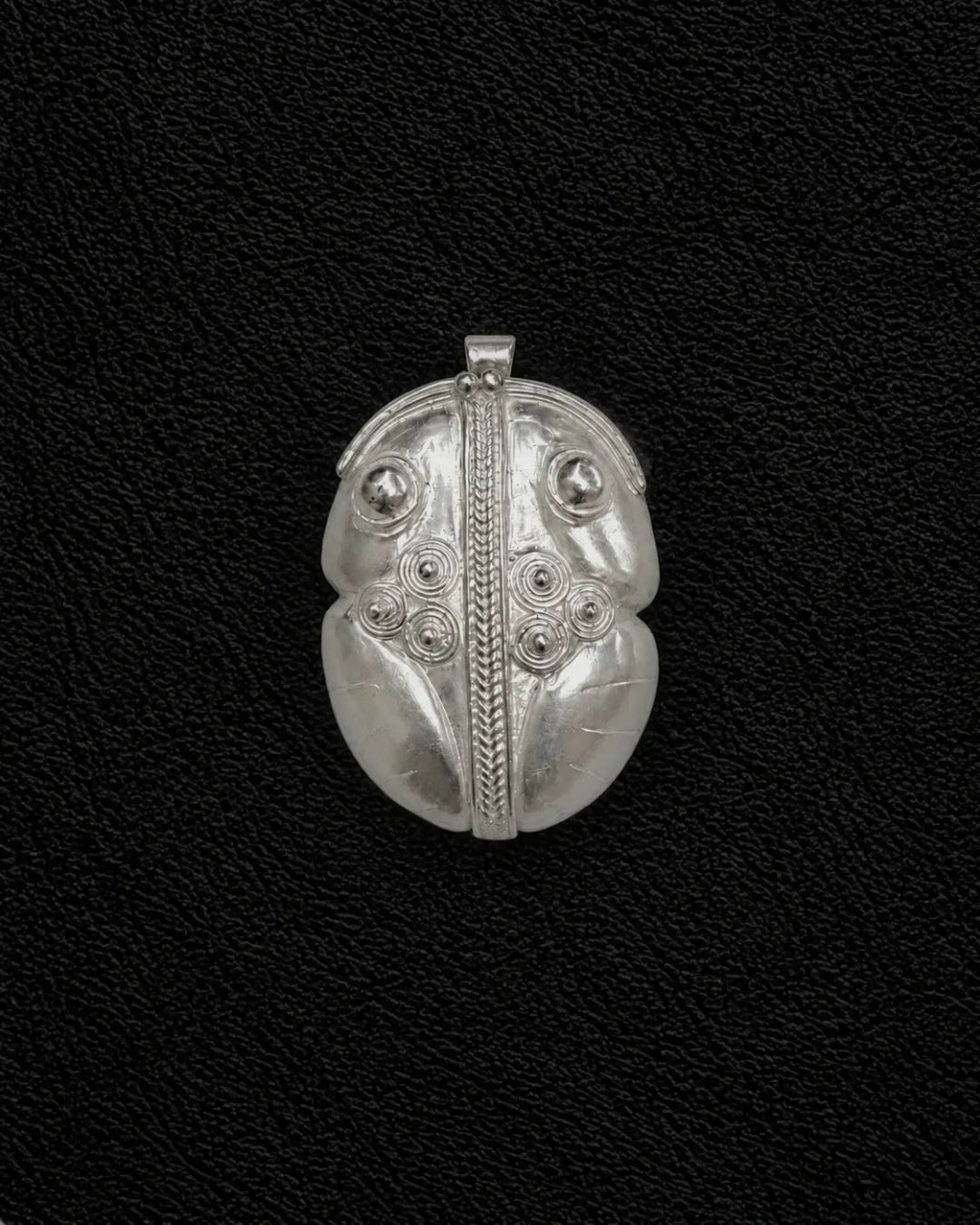 A silver pendant with intricate tribal-style engraved patterns, including a central vertical line, circular details, and leaf-like sections, displayed on a textured black background.