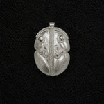 A silver pendant with intricate tribal-style engraved patterns, including a central vertical line, circular details, and leaf-like sections, displayed on a textured black background.