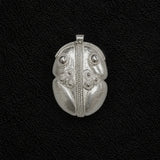 A silver pendant with intricate tribal-style engraved patterns, including a central vertical line, circular details, and leaf-like sections, displayed on a textured black background.