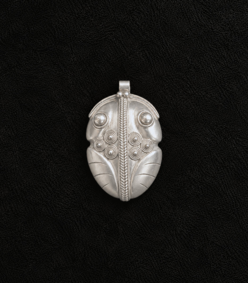 A silver pendant with intricate tribal-style engraved patterns, including a central vertical line, circular details, and leaf-like sections, displayed on a textured black background.