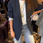 A person wearing an oversized dark gray blazer over a white T-shirt and light-wash jeans, styled with layered gold necklaces in a boutique setting.