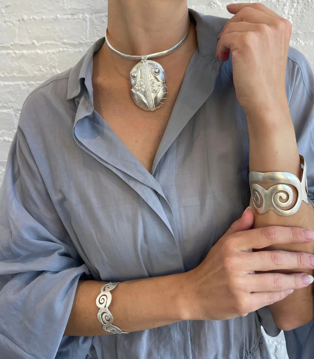 A person wearing a silver-toned jewelry set, including a leaf-shaped pendant necklace, a matching cuff bracelet, and a spiral-patterned bangle, styled with a light blue linen shirt against a white brick wall.