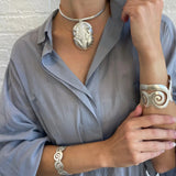 A person wearing a silver-toned jewelry set, including a leaf-shaped pendant necklace, a matching cuff bracelet, and a spiral-patterned bangle, styled with a light blue linen shirt against a white brick wall.