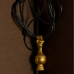 A gold pendant necklace with a decorative vase-like charm on a black cord, photographed against a dark brown background.