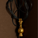 A gold pendant necklace with a decorative vase-like charm on a black cord, photographed against a dark brown background.