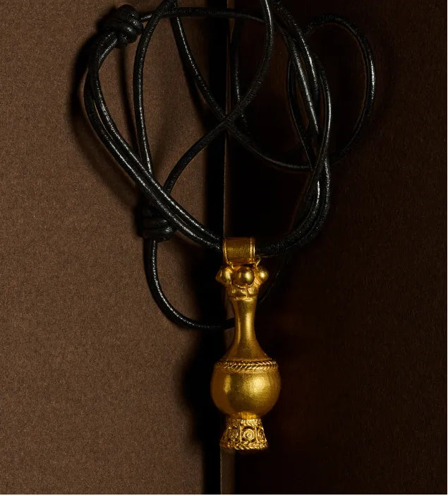A gold pendant necklace with a decorative vase-like charm on a black cord, photographed against a dark brown background.