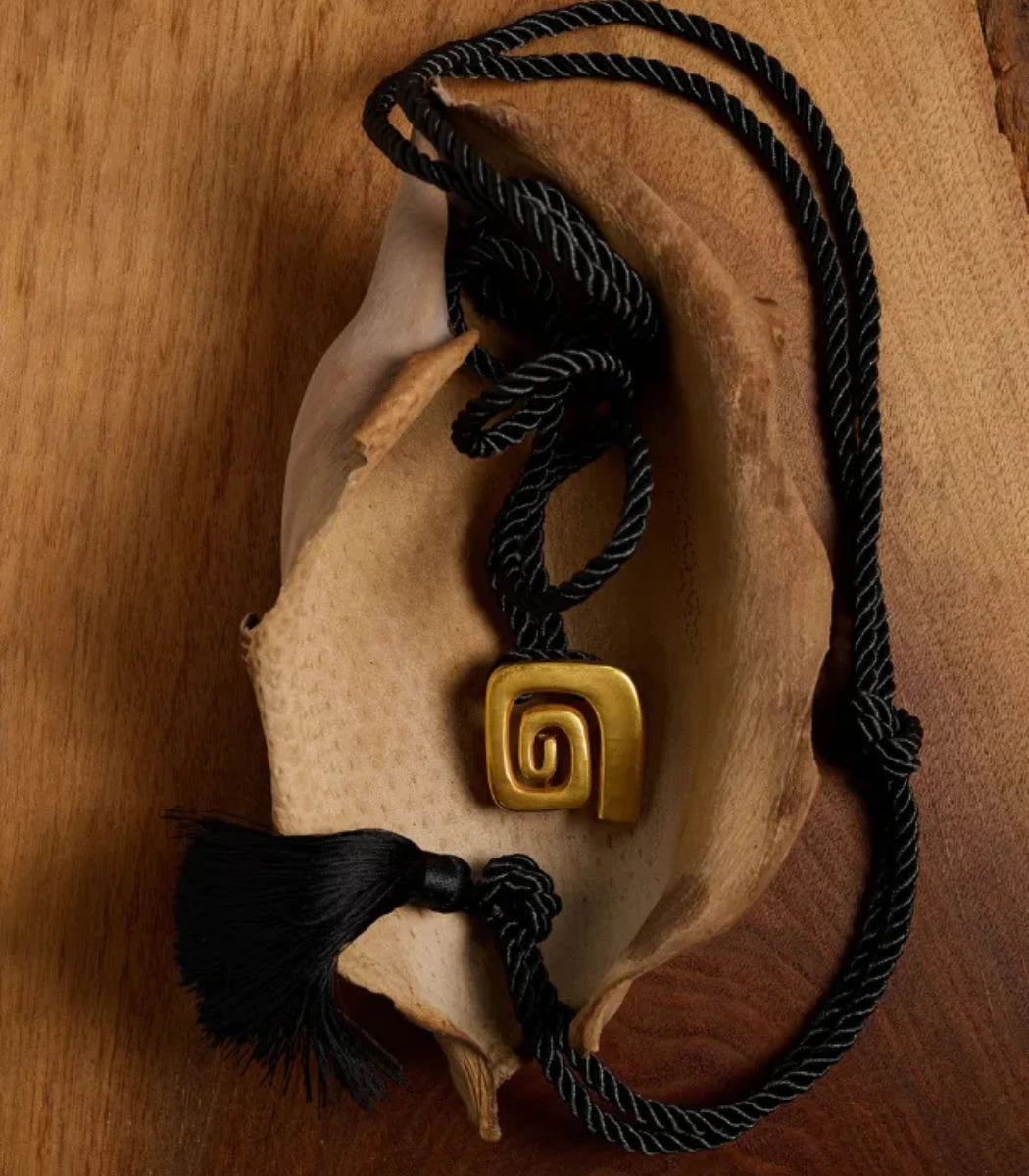 A black braided cord necklace with a gold spiral pendant, displayed on a dried leaf over a wooden surface.