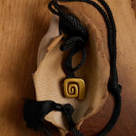 A black braided cord necklace with a gold spiral pendant, displayed on a dried leaf over a wooden surface.