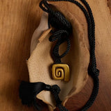 A black braided cord necklace with a gold spiral pendant, displayed on a dried leaf over a wooden surface.